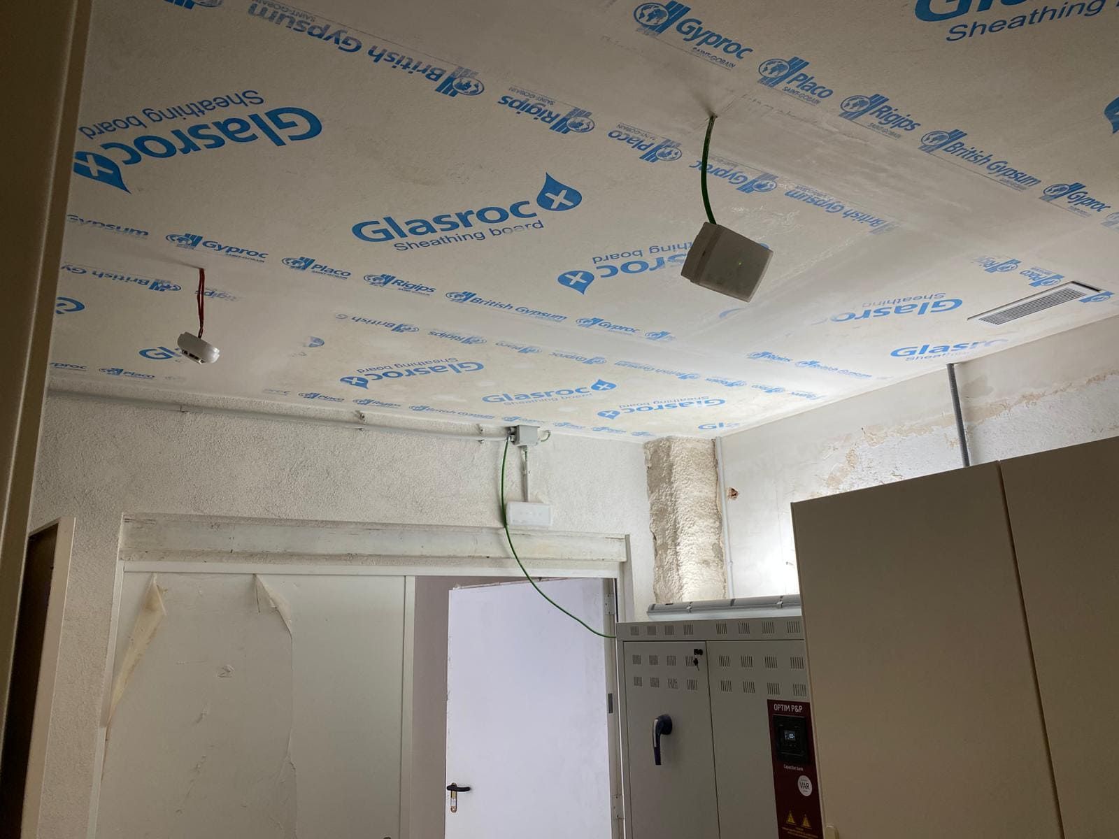 Techo con paneles Glasroc, caja eléctrica, cables expuestos, habitación sin terminar, puerta blanca.