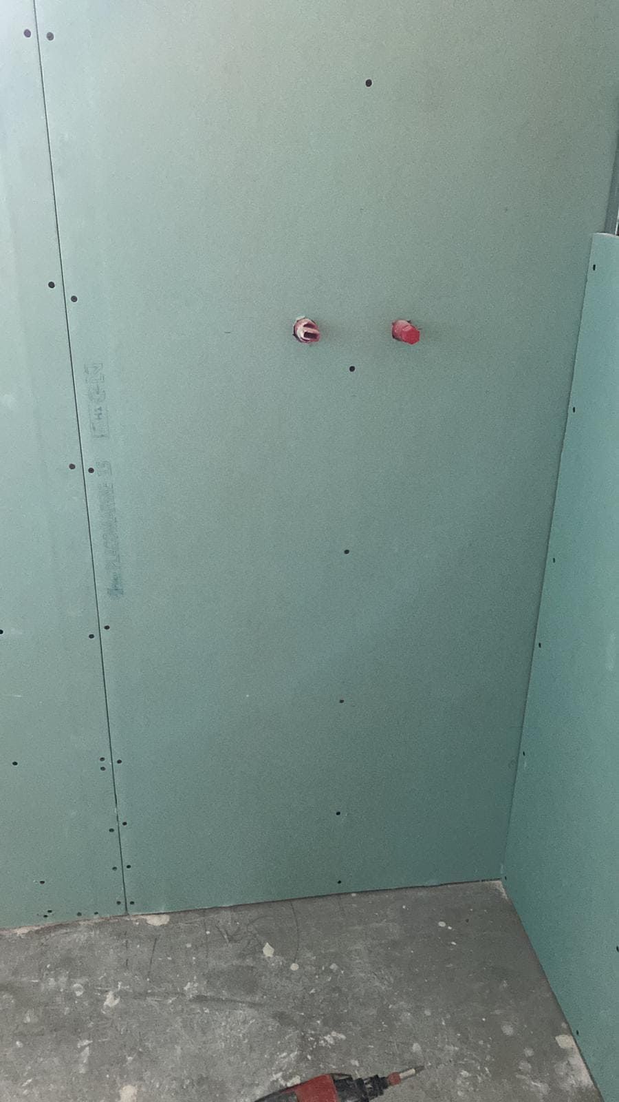 Panel de yeso verde en un entorno de construcción con dos tuberías con tapas rojas visibles.