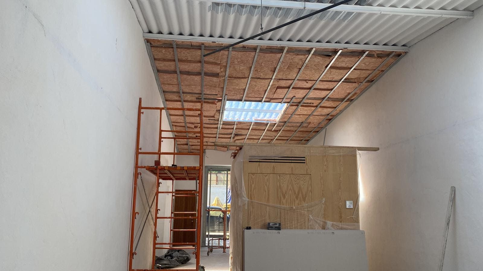 Habitación interior en construcción con andamios, techo dañado y claraboya. Paredes blancas.