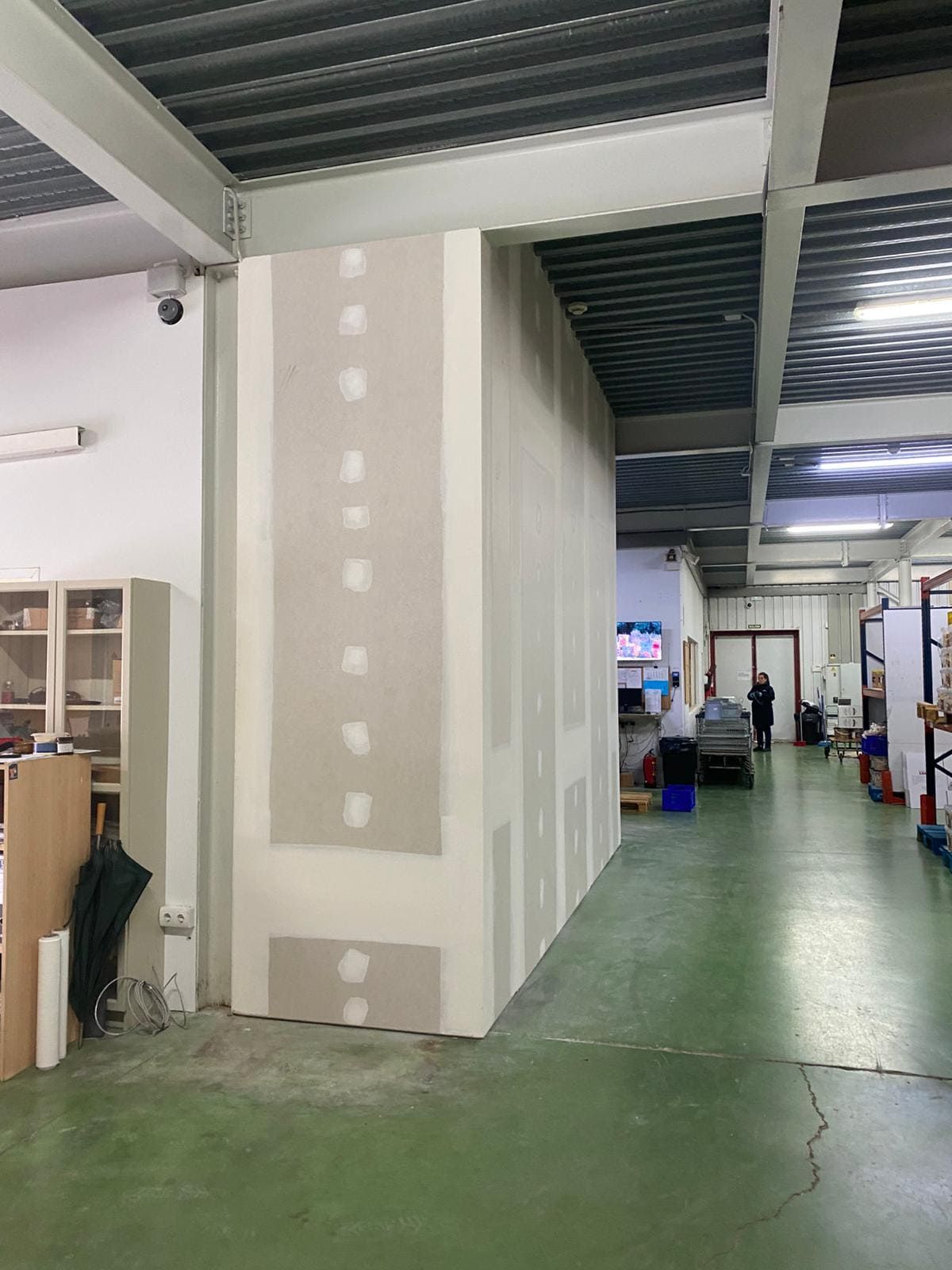 Vista interior de fábrica con partición de yeso; piso verde, techo de metal, gente en la distancia.