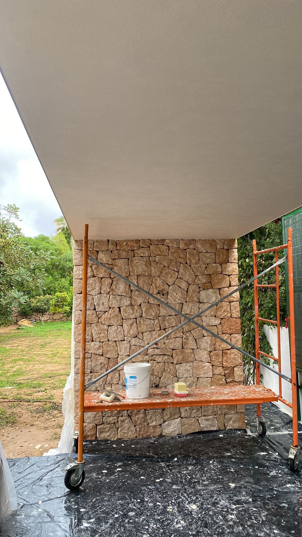 Andamio frente a un muro de piedra, bajo un techo blanco. Un cubo y herramientas descansan sobre la plataforma del andamio.