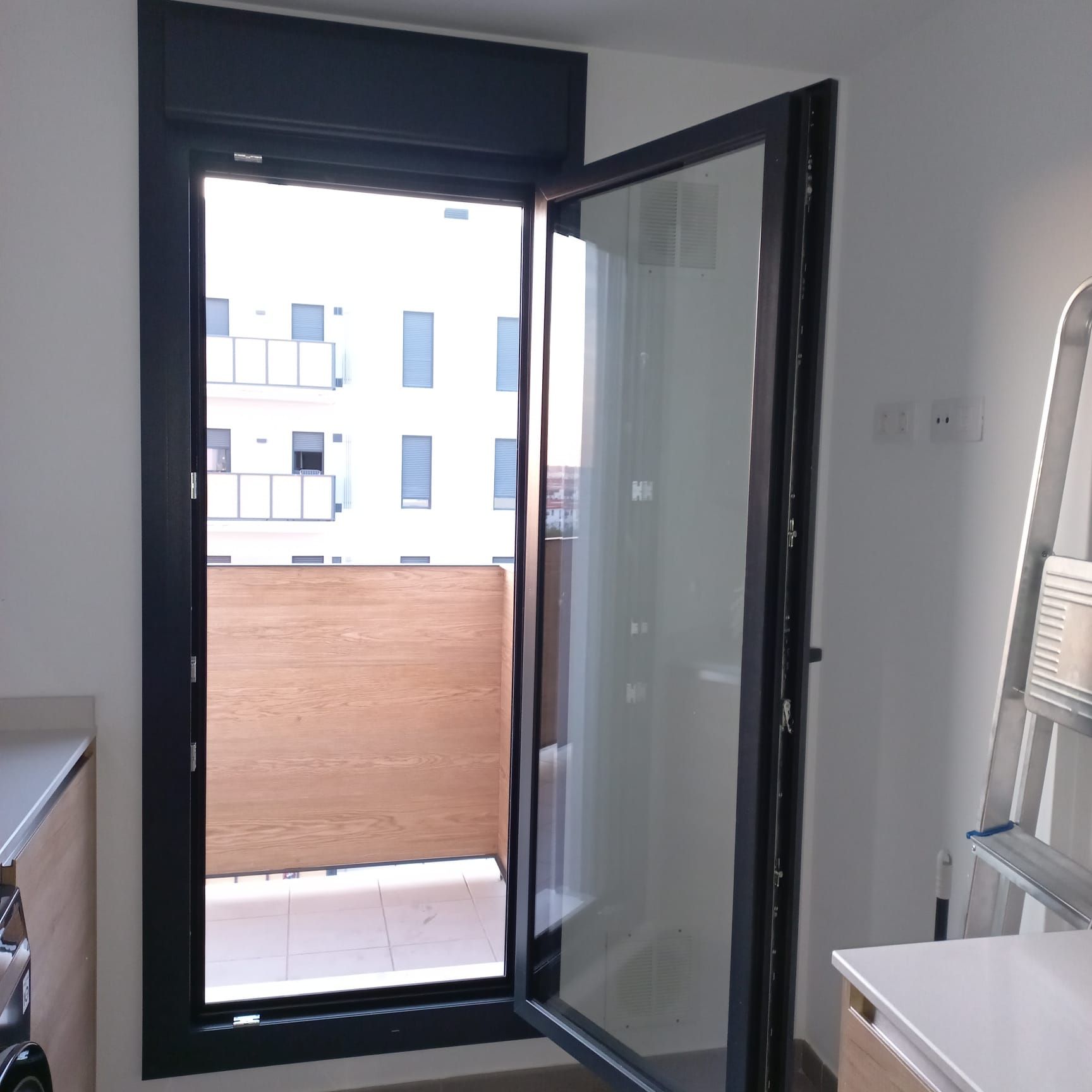 Puerta de cristal que da a un balcón con vista al edificio, con marco oscuro. Escalera, armario y pared en la habitación.