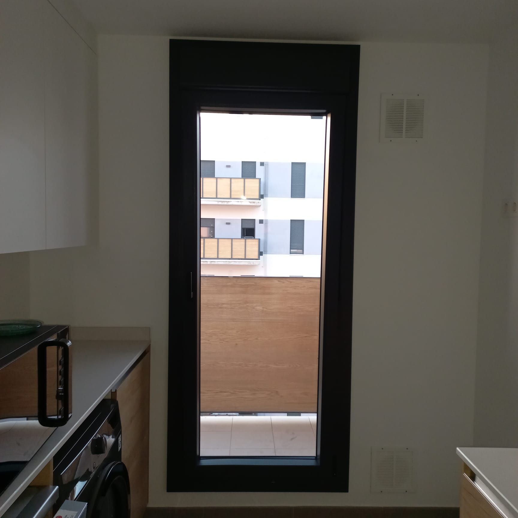 Una ventana de cocina con marco negro, con vista a un edificio, paredes blancas y una encimera.