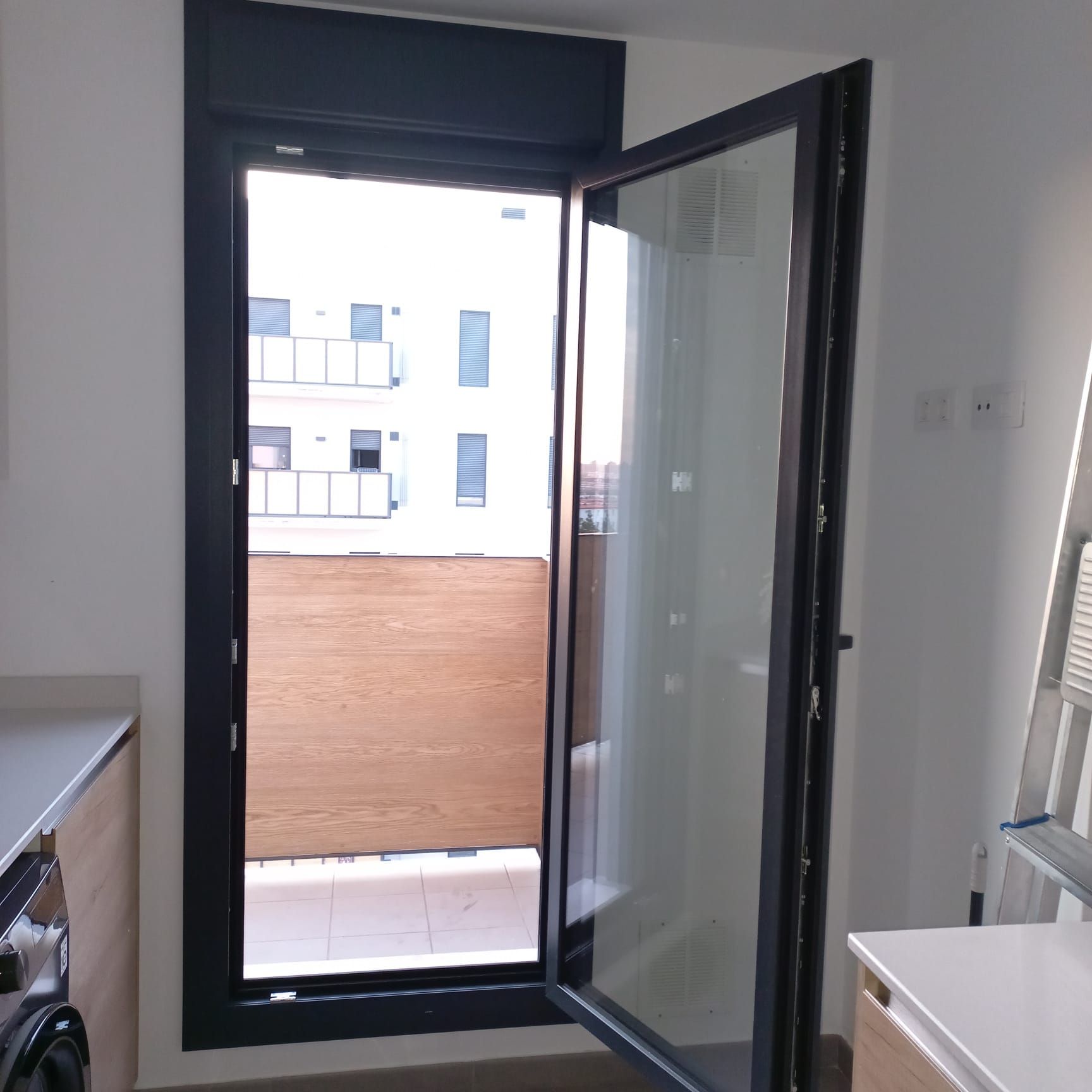 Puerta de cristal abierta a un balcón. Marco negro, puerta de madera debajo, edificio blanco afuera.