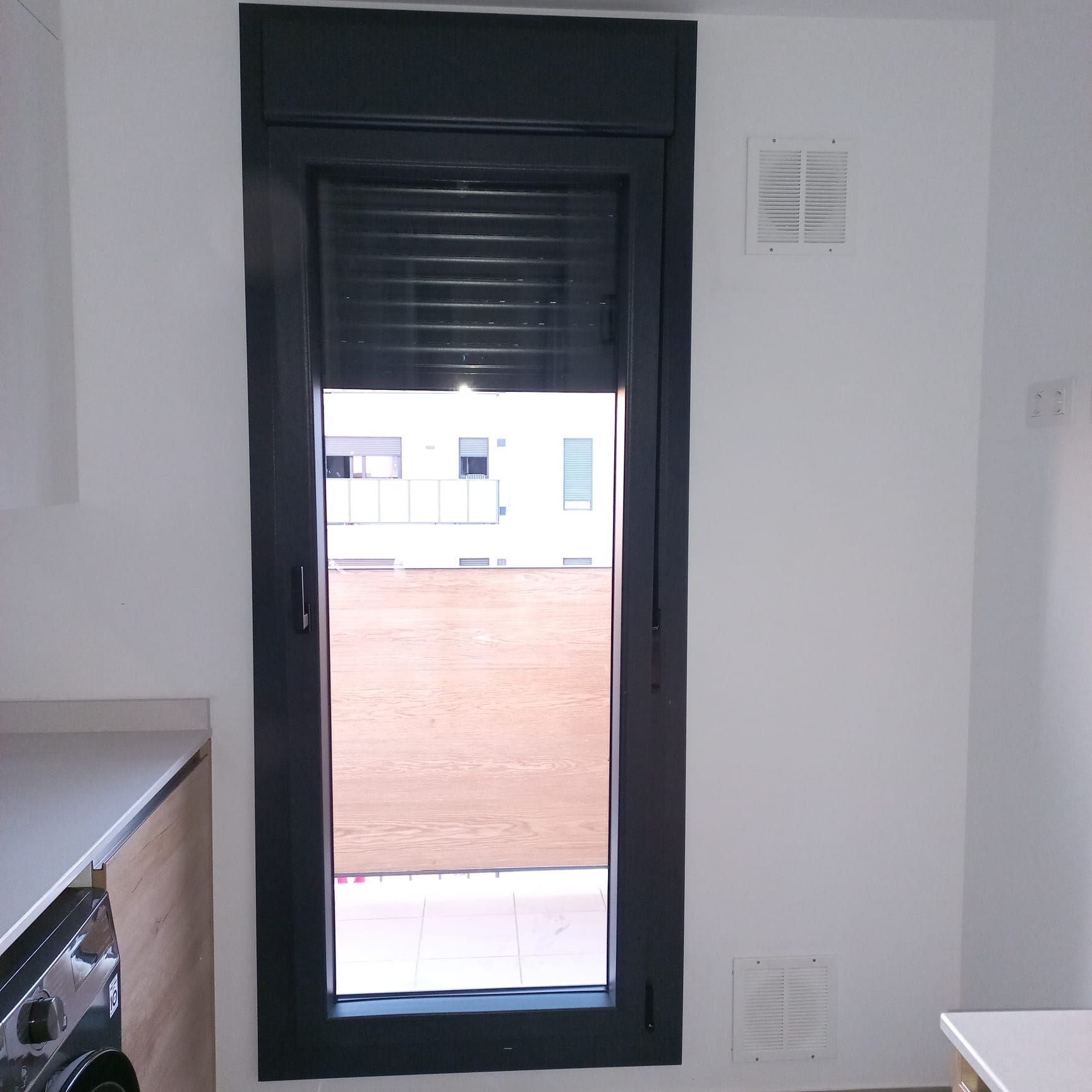 Ventana de marco oscuro con persianas cerradas. Paredes blancas y una rejilla de ventilación visible.