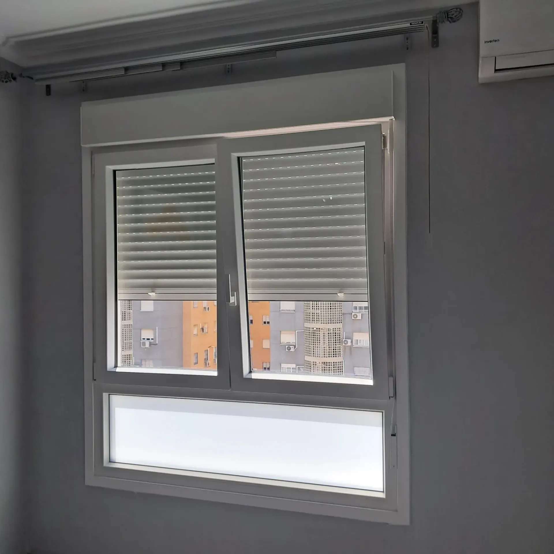 Ventana blanca con persianas cerradas, que oculta parcialmente la vista de los edificios.