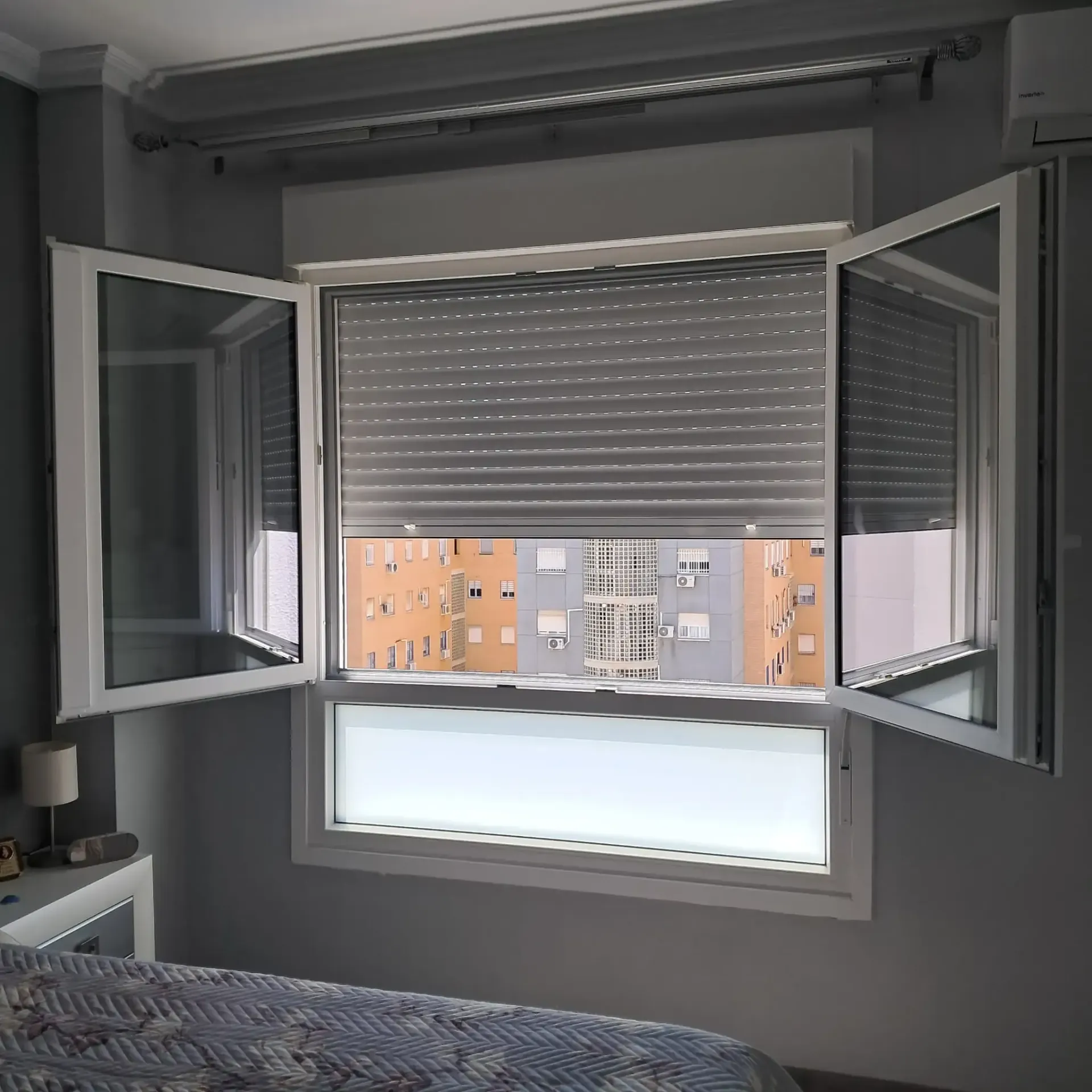 Una ventana con persianas grises ofrece vistas a la ciudad. La habitación tiene paredes grises y una cama.