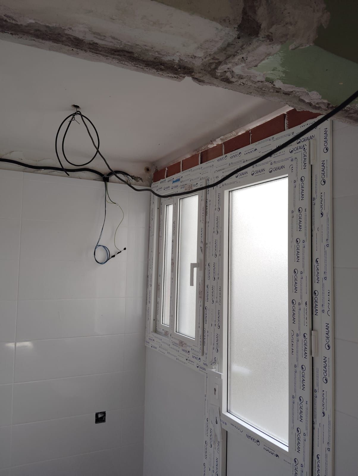 Vista interior con paredes de azulejos blancos, un marco de ventana y puerta, cables expuestos.