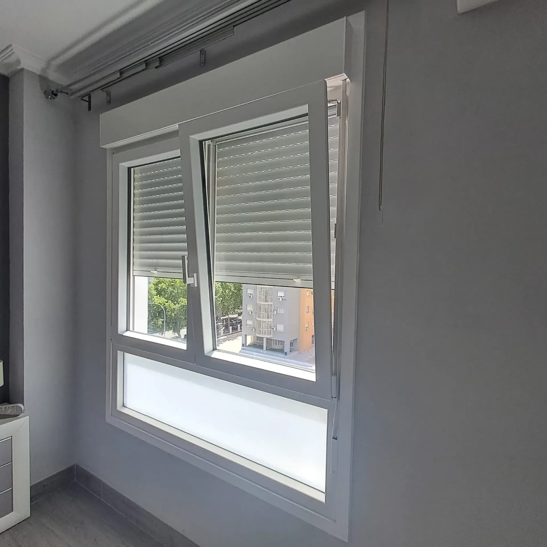 Ventana con persianas blancas que cubren parcialmente la vista de los edificios y la vegetación.