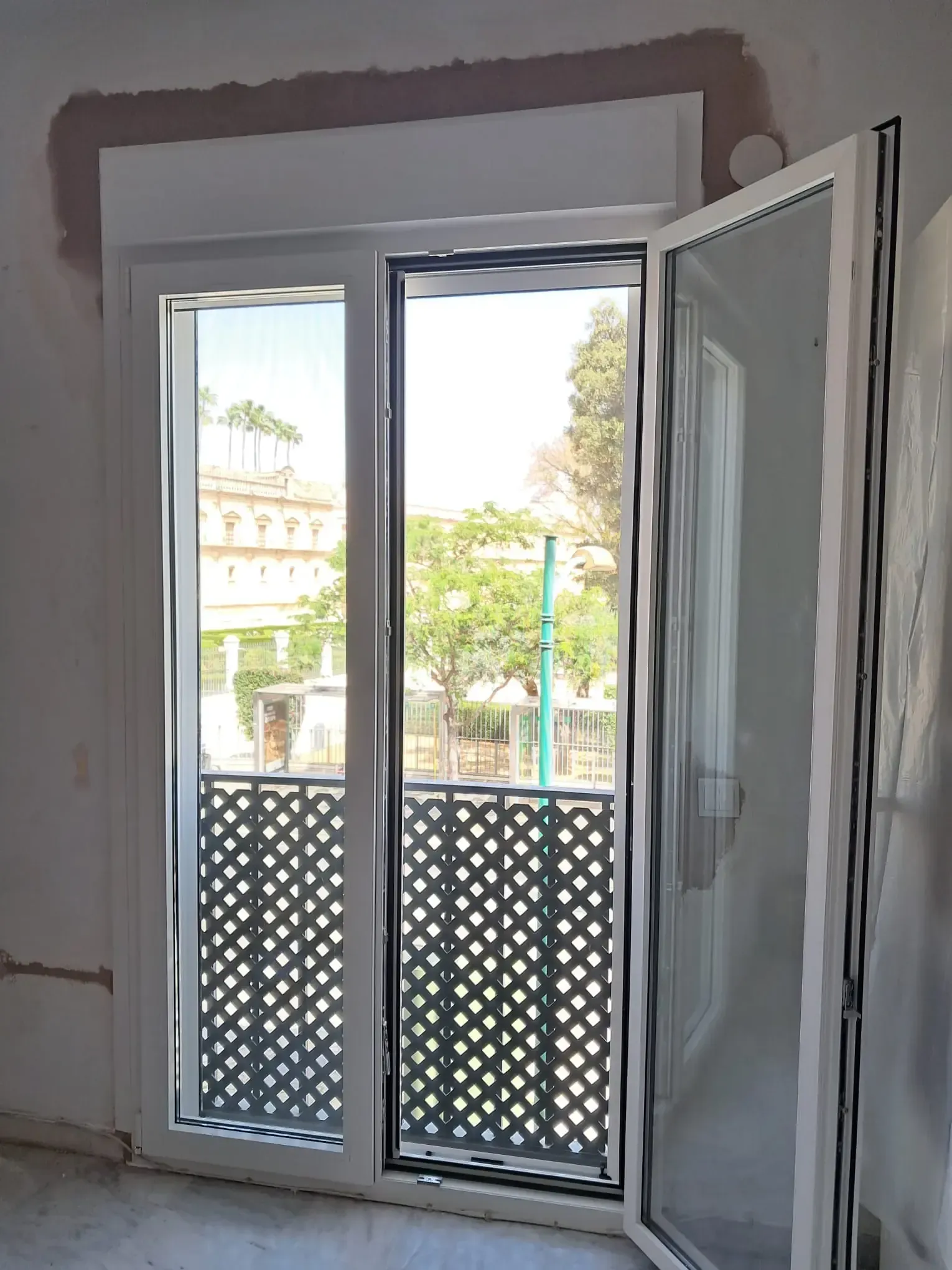 Puerta y ventana blancas abiertas que dan a una calle con un balcón negro decorativo.