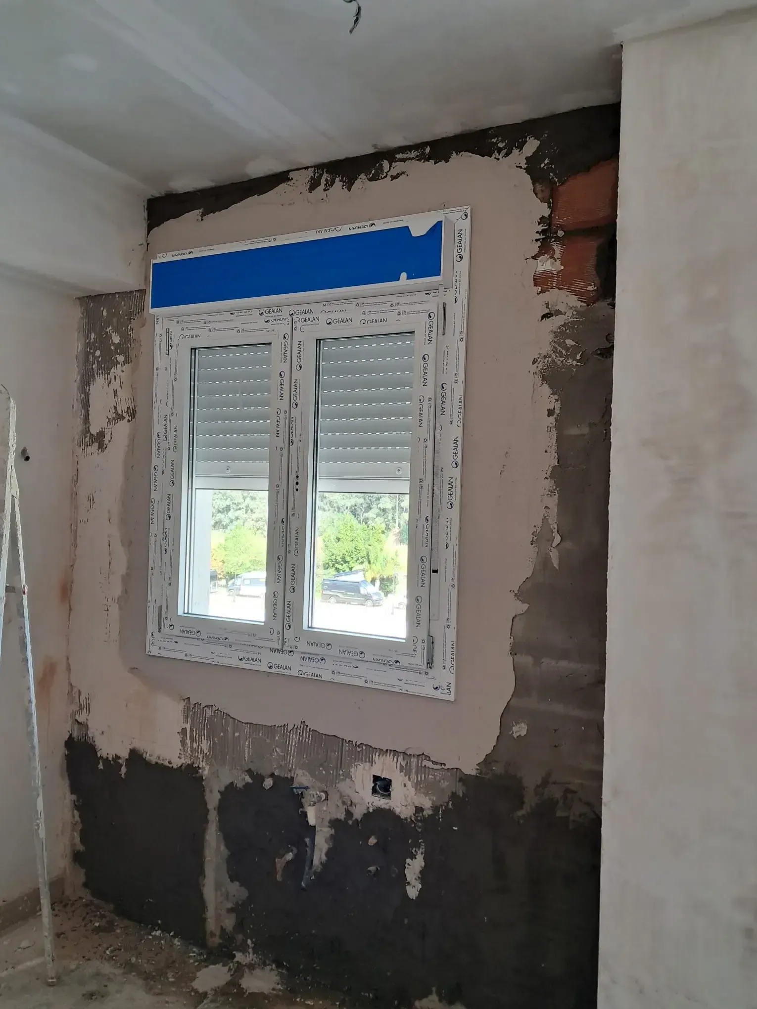 Ventana en una habitación en construcción con marco blanco, cinta azul y ladrillo visto.