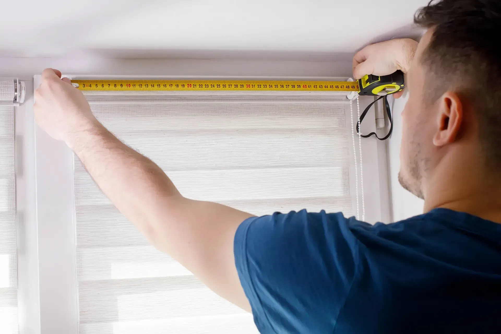 Persona midiendo una ventana para persianas con una cinta métrica amarilla.