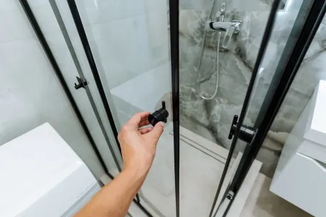 Mano abriendo una puerta de ducha con marco negro con una pared de azulejos de mármol gris en el fondo.