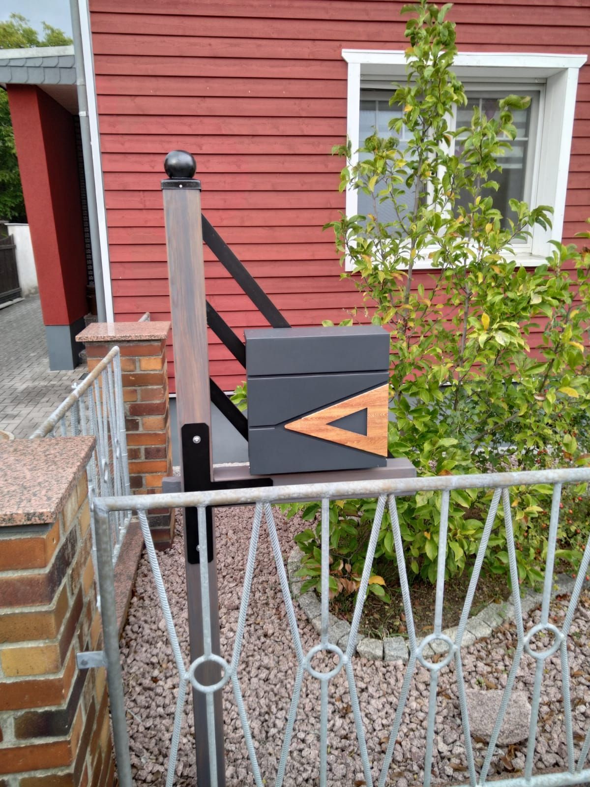 Ein Briefkasten steht neben einem Zaun vor einem roten Haus.