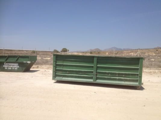 Un contenedor de basura verde está en el medio de un campo de tierra.