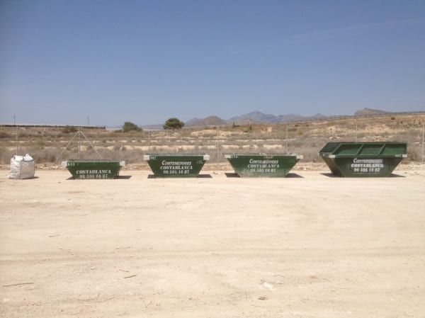 Tres contenedores de basura verdes están alineados en un campo de tierra.