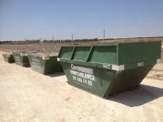 Una fila de contenedores de basura verdes se encuentran encima de un campo de tierra.