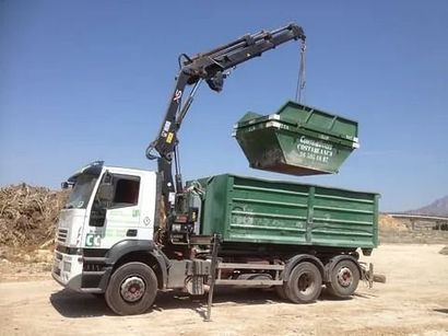 Un camión volquete está siendo cargado con un contenedor verde mediante una grúa.