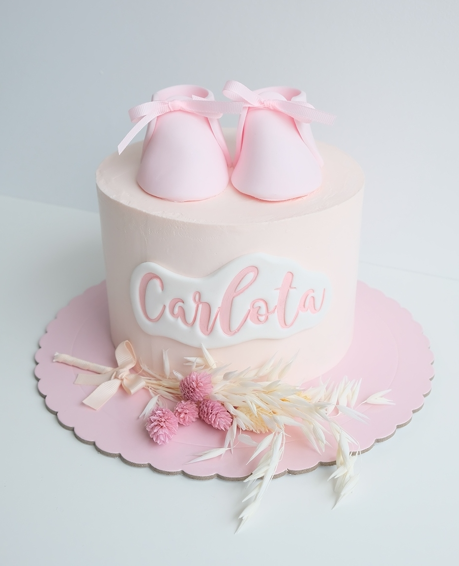 Un pastel rosa con un par de zapatitos de bebé rosas encima. de nombre carlota
