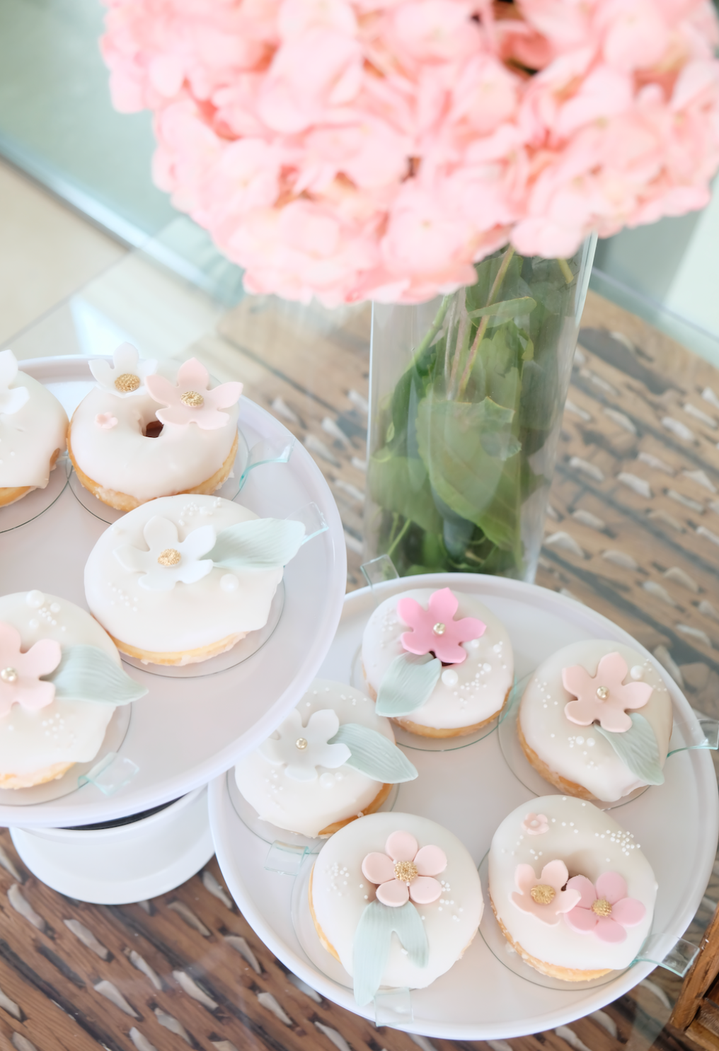 Dos platos de donas con flores rosas sobre ellos están en una mesa al lado de un jarrón de flores rosas.