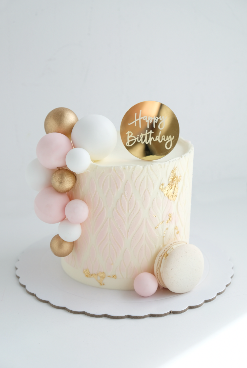 Un pastel de cumpleaños blanco con globos rosas y dorados y macarons encima.