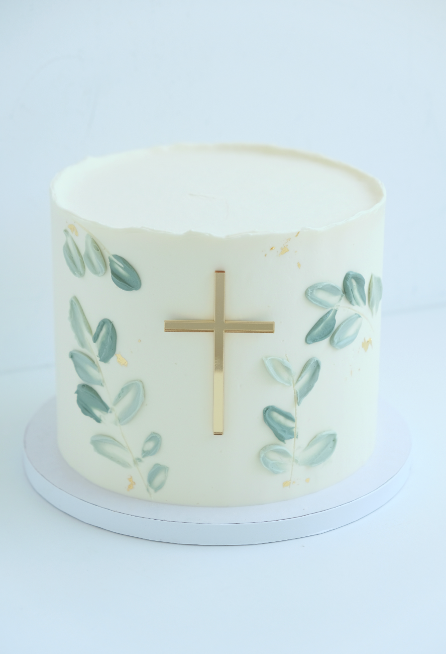 Un pastel blanco con una cruz dorada y hojas verdes encima.