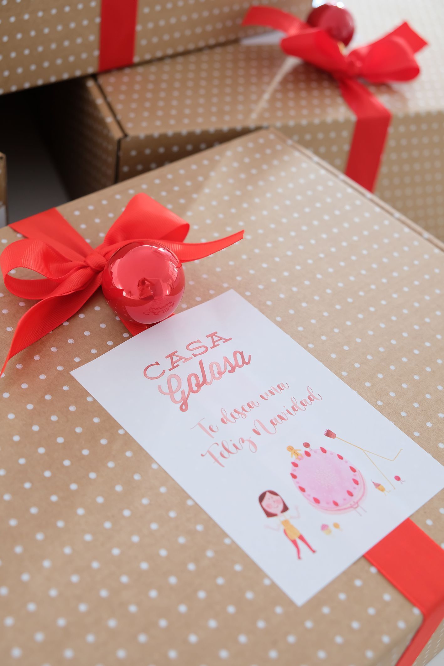 Caja de carton con moño rojo y bola y tarjeta casa golosa