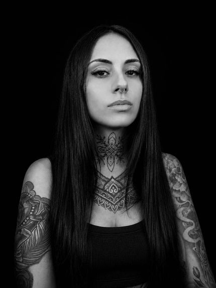 Retrato de una persona tatuada con cabello largo y oscuro sobre un fondo negro, mirando al frente con expresión neutra.