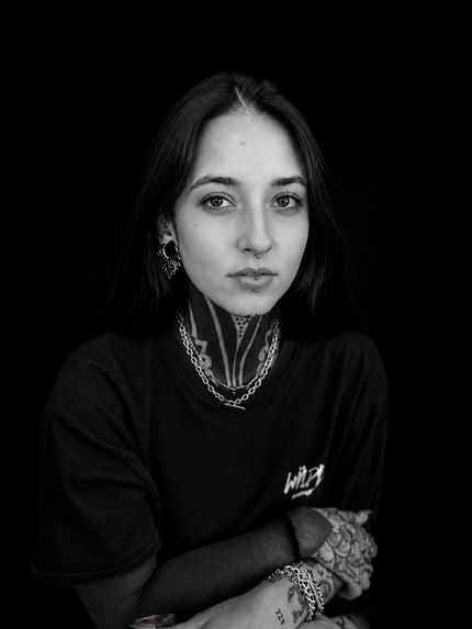 Retrato en blanco y negro de una persona con cabello oscuro, pendientes y collares superpuestos, mirando a la cámara.
