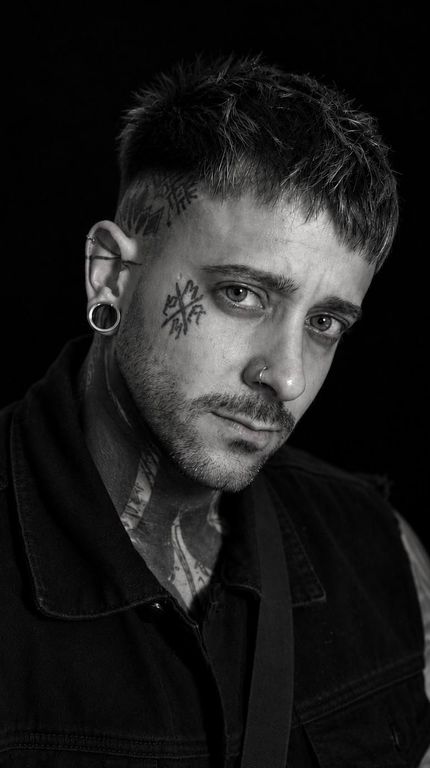 Retrato de una persona tatuada con cabello largo y oscuro sobre un fondo negro, mirando al frente con expresión neutra.