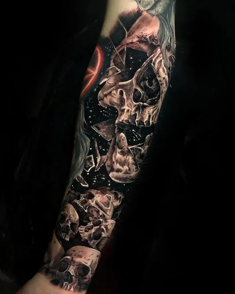 Brazo cubierto por un tatuaje oscuro y realista de una calavera y un rostro sobre un fondo negro.