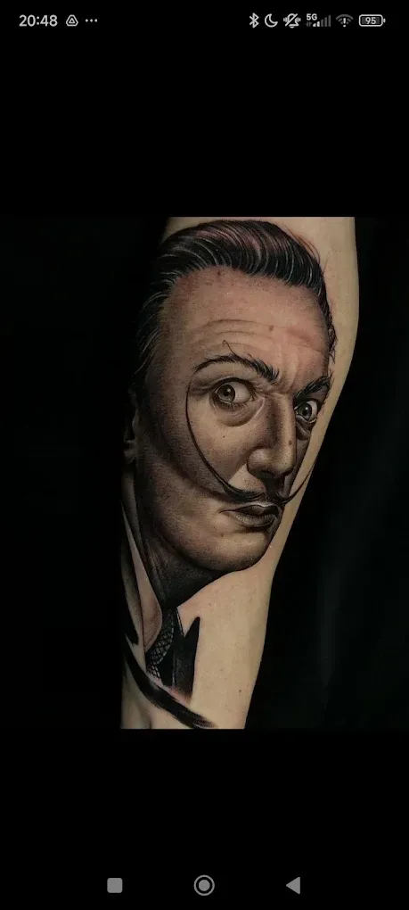 Tatuaje de retrato de una cara sonriente en el brazo de una persona, realizado con tinta oscura y sombreado detallado.