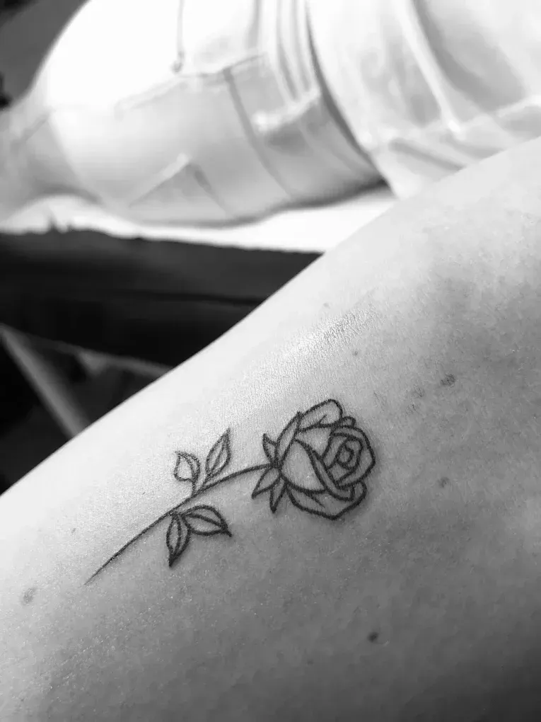 Primer plano en blanco y negro de un tatuaje de una rosa en el antebrazo de una persona que descansa sobre una silla o mesa.