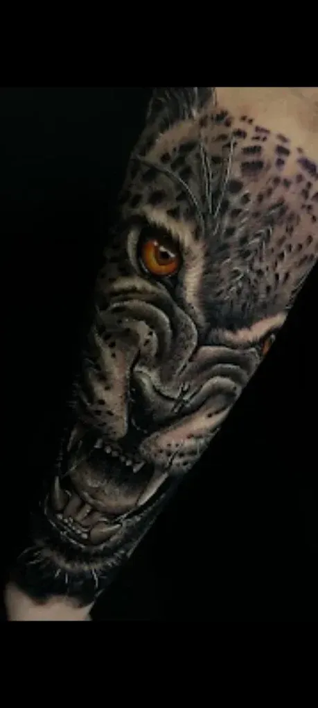 Tatuaje detallado de la cara de un tigre en el antebrazo, con un ojo naranja realista y un patrón de manchas en el pelaje.