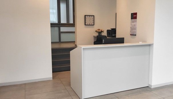 Weißer Empfangstresen in einem Büro. An der Wand hängen eine Uhr und ein Schild; eine kleine Treppe und ein Fenster sind zu sehen.