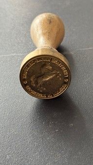 Holzstempel mit einem runden Stempelkopf aus Messing. Der Kopf trägt ein geprägtes Bild und einen Text.