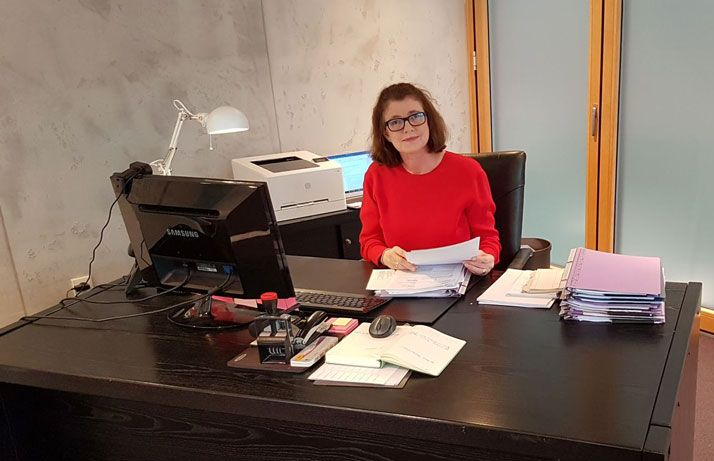 Eine Frau in einem roten Pullover sitzt an einem Schreibtisch mit Papieren und schaut in die Kamera.