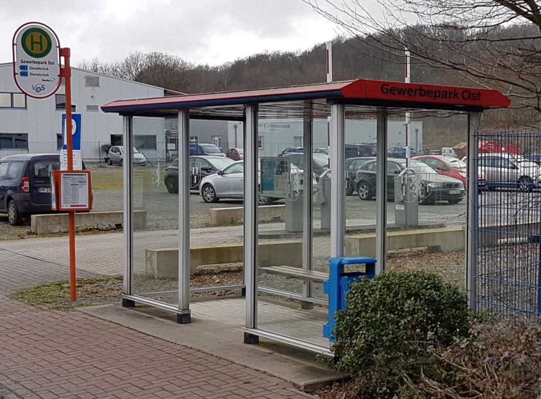 Bushaltestelle mit gläsernem Wartehäuschen, Schild und Sitzgelegenheiten. Im Hintergrund sind Autos geparkt.