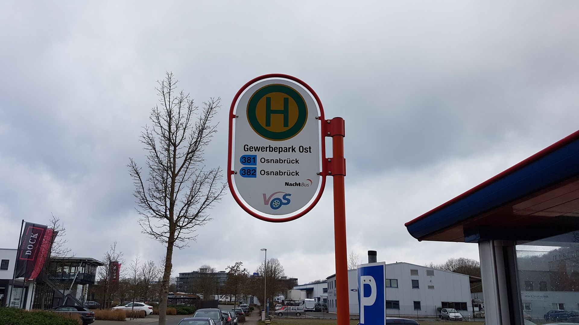 Bushaltestellenschild mit grünem „H“ in einem weißen ovalen Rahmen an einem roten Mast unter bewölktem Himmel.
