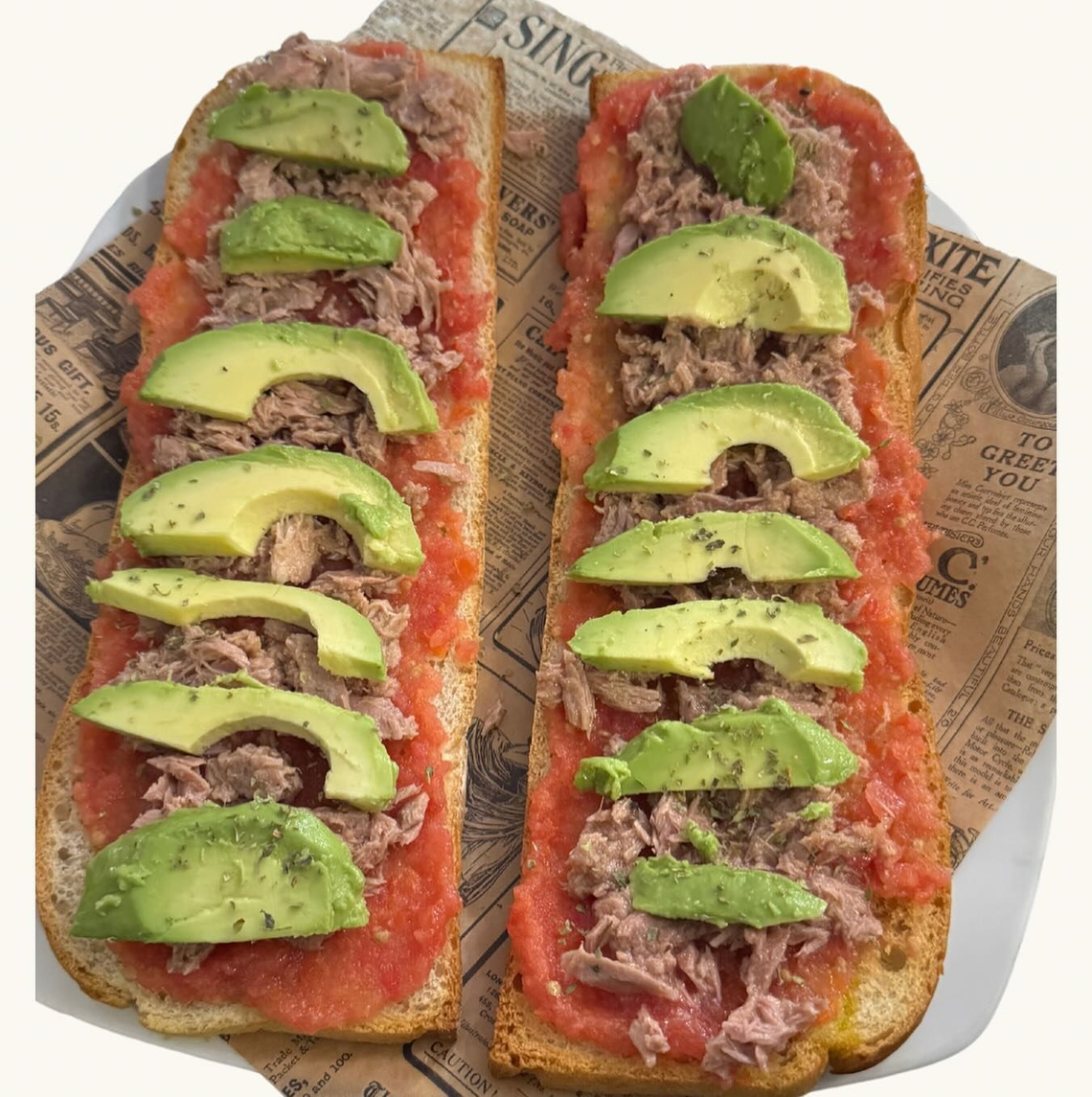 Dos mitades de tostada de aguacate sobre papel de periódico, adornadas con verduras y crema roja, sobre un fondo claro con detalles verdes.