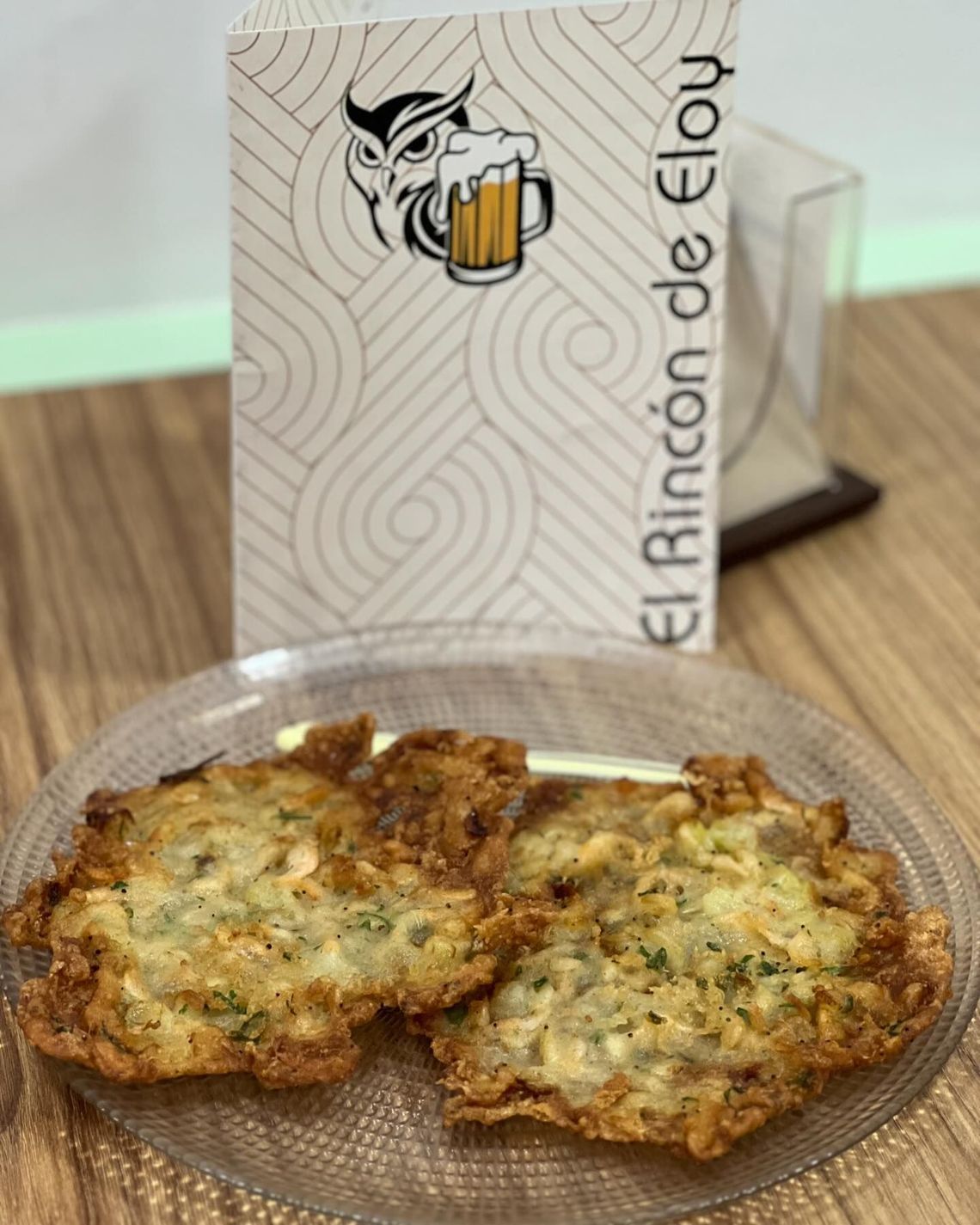 Tres hamburguesas doradas fritas en un plato transparente junto a una bolsa con la marca “El Rincón de ELO” sobre una mesa.