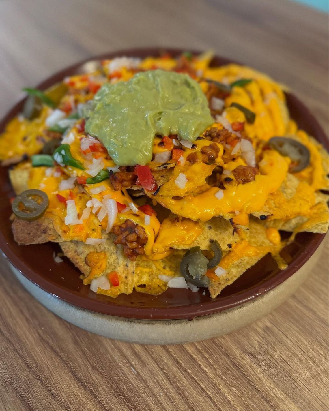 Plato de nachos cubiertos con guacamole, queso, jalapeños y hojuelas de pimiento rojo sobre una mesa de madera.