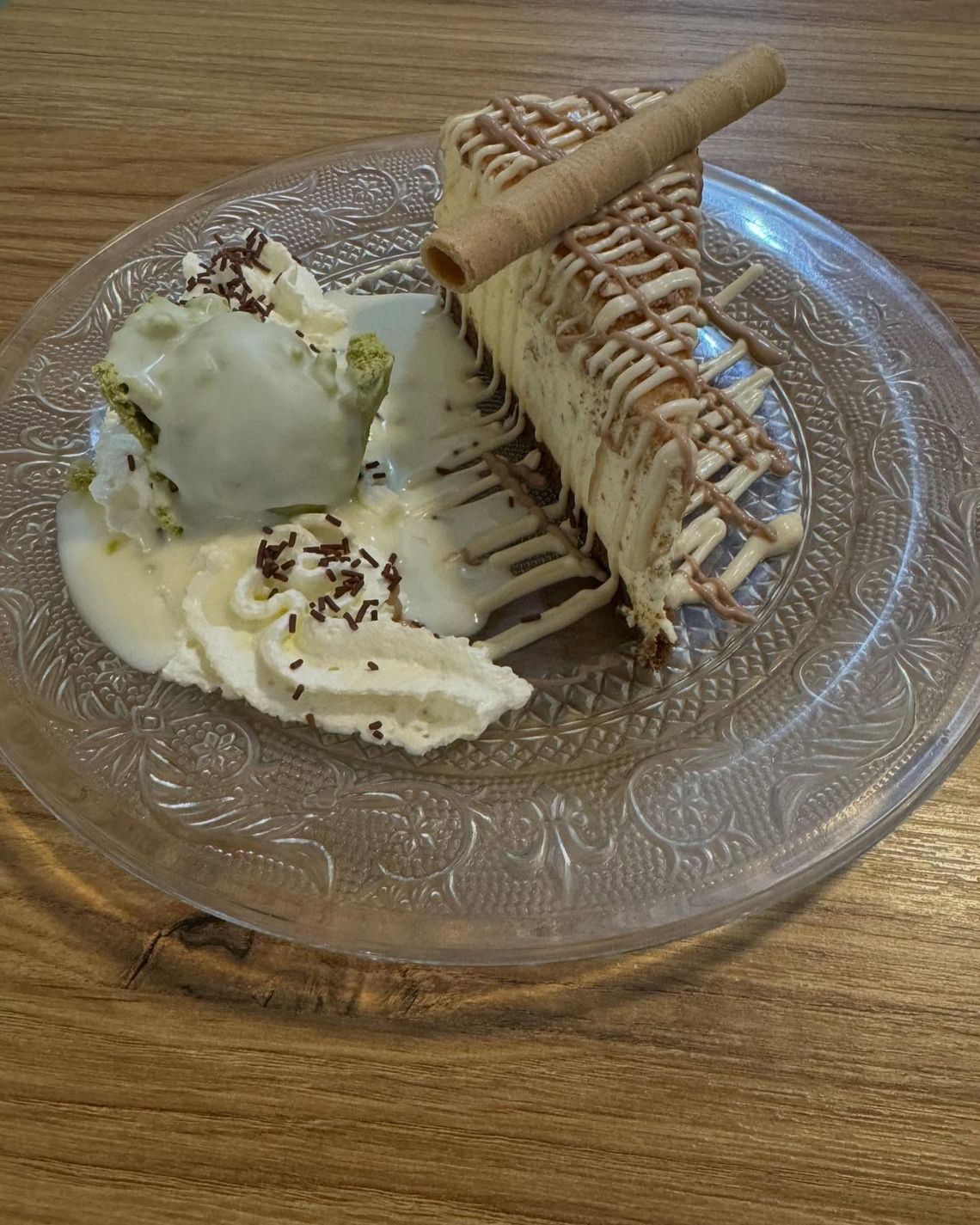 Porción de postre con barquillo de chocolate y helado en un plato de cristal, sobre una mesa de madera.