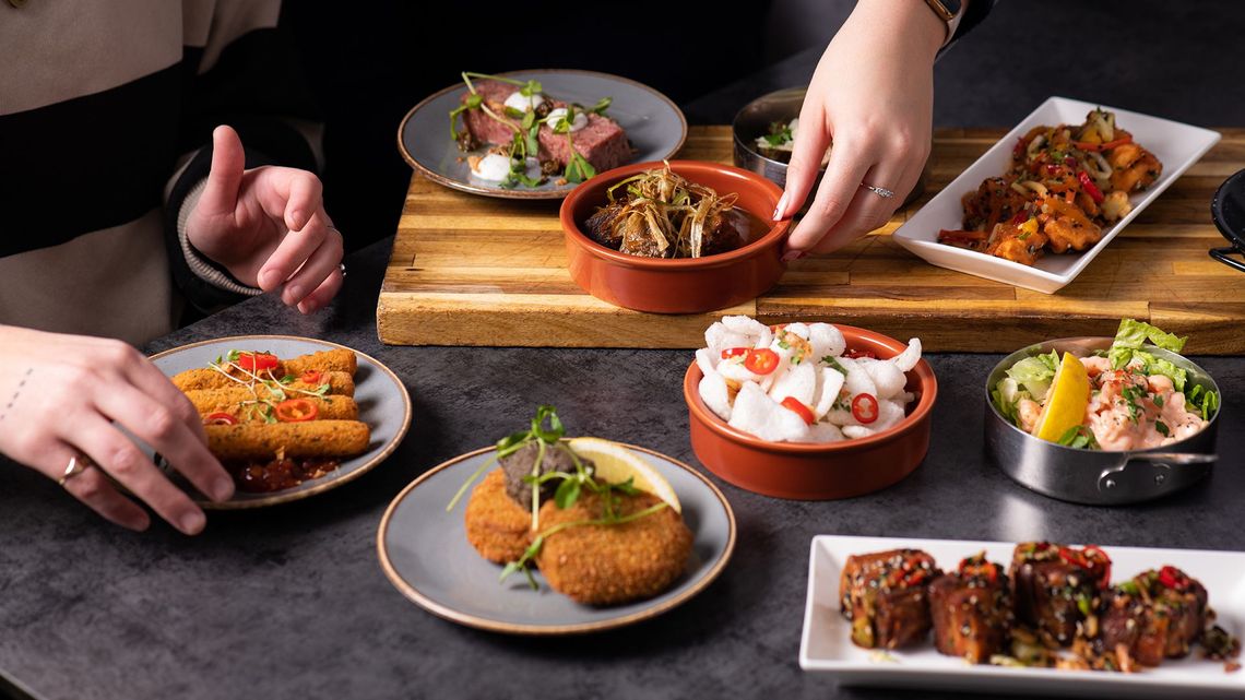Variedad de aperitivos servidos en platos sobre una mesa oscura, con manos que se extienden para coger los platos durante una comida compartida.