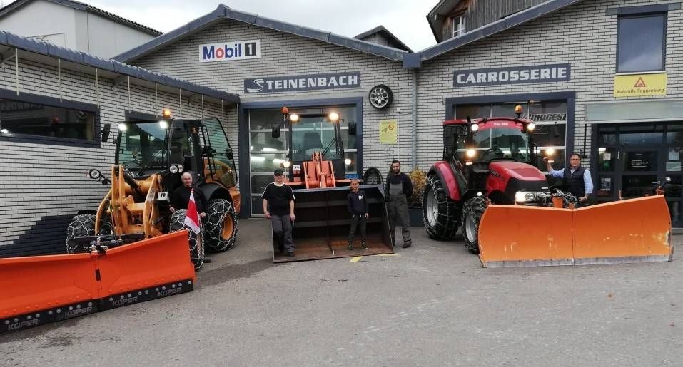 Ein Geschäft mit Traktoren und Schneepflügen davor, mehrere Leute posieren, das Mobil 1-Schild ist zu sehen.