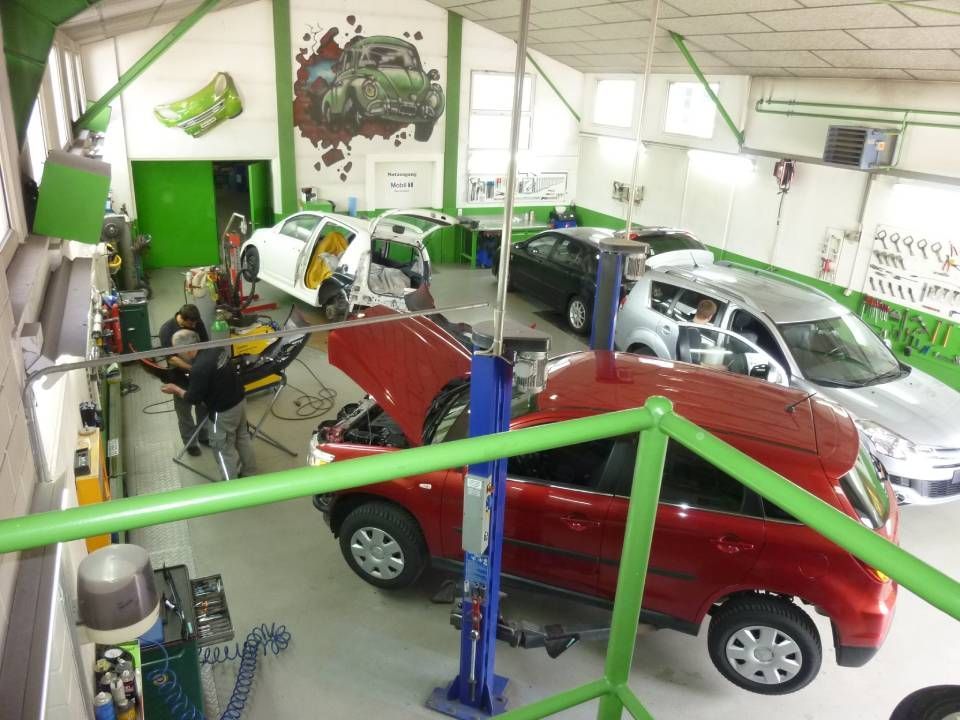 Autos in einer geschäftigen Autowerkstatt. Ein Wagen steht auf der Hebebühne, andere befinden sich in unterschiedlichen Reparaturstadien. Grün-weisse Dekoration.