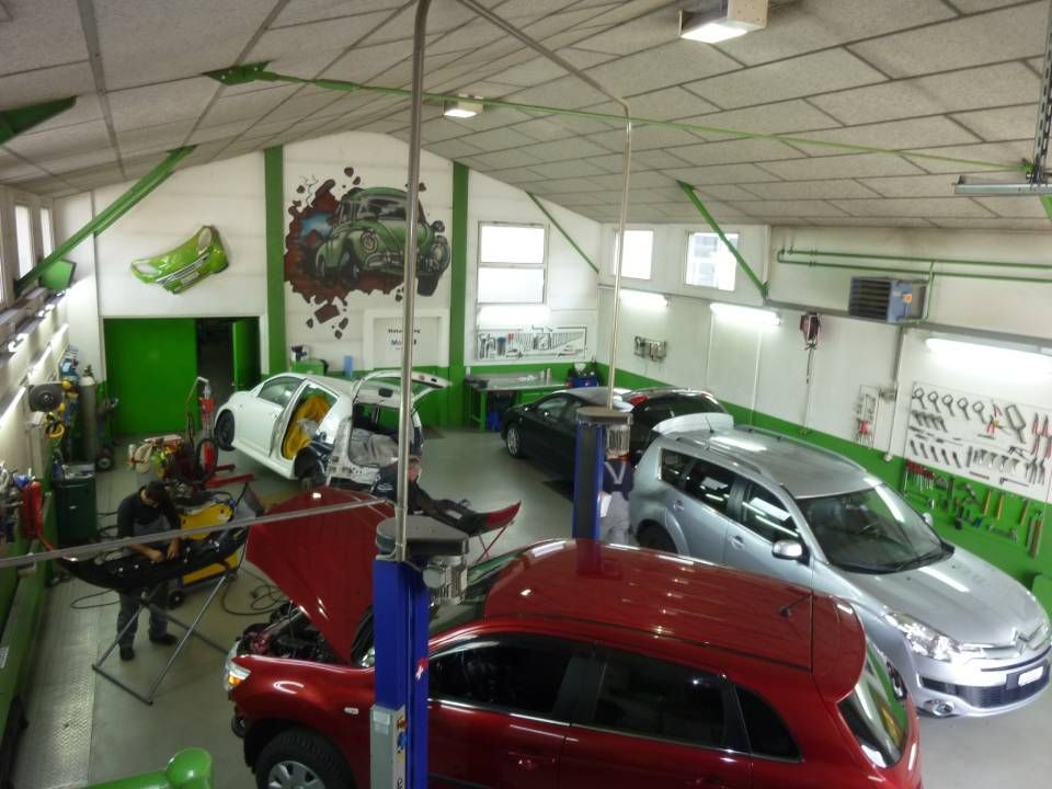 Innenansicht einer Autowerkstatt mit Fahrzeugen in unterschiedlichen Reparaturzuständen. Grün-weisse Dekoration, Deckenbeleuchtung.