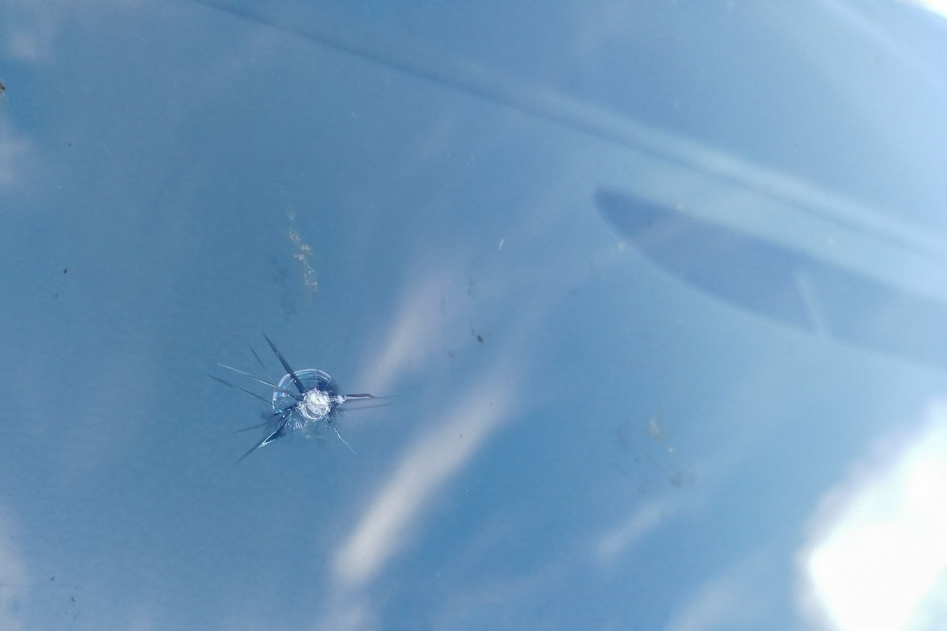 Pare-brise de voiture fissuré, présentant un impact en forme d'œil-de-bœuf et des fissures rayonnant vers l'extérieur, sur fond de ciel bleu.