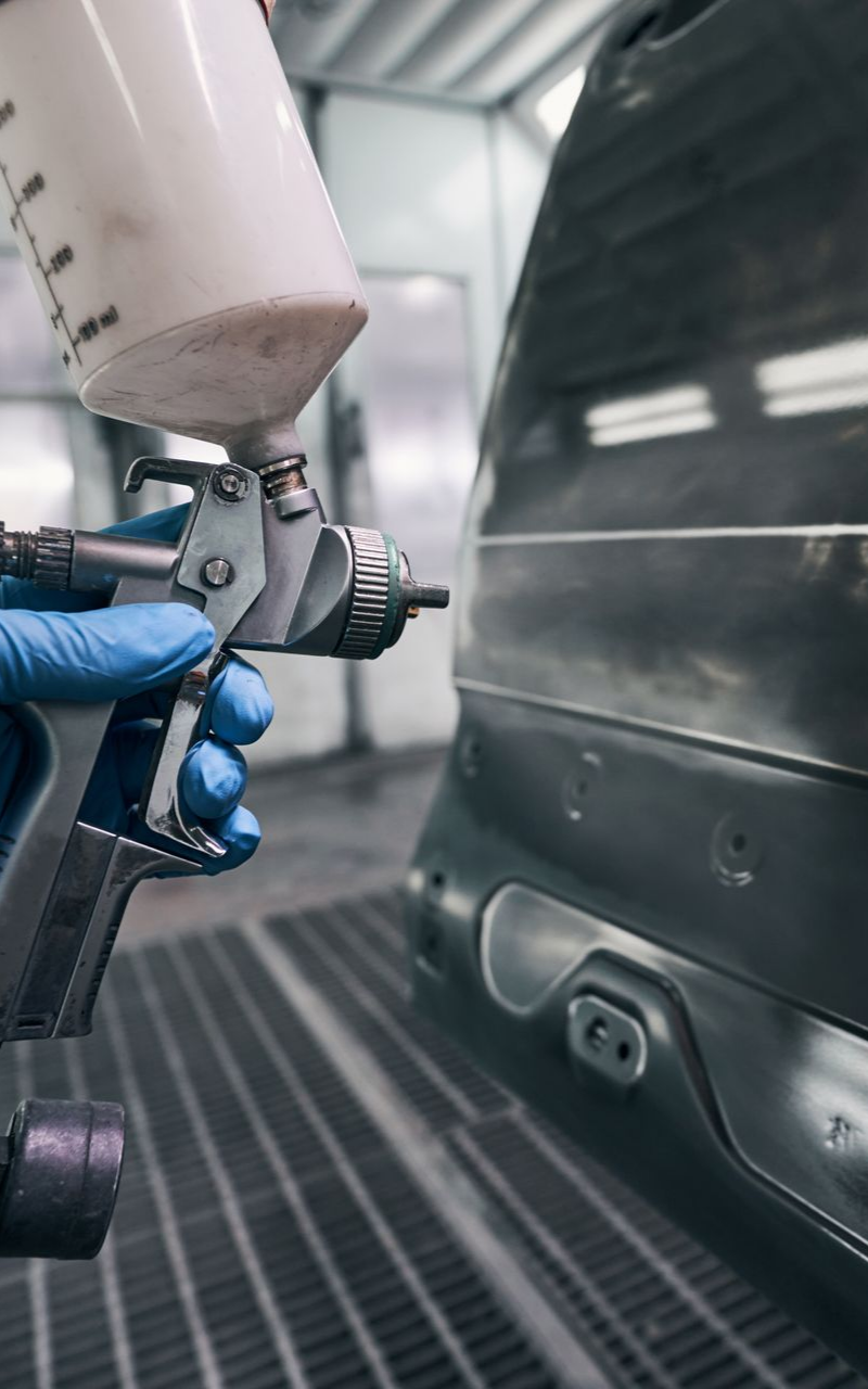 Une personne portant des gants bleus utilise un pistolet à peinture pour peindre un panneau de voiture dans une cabine de peinture.