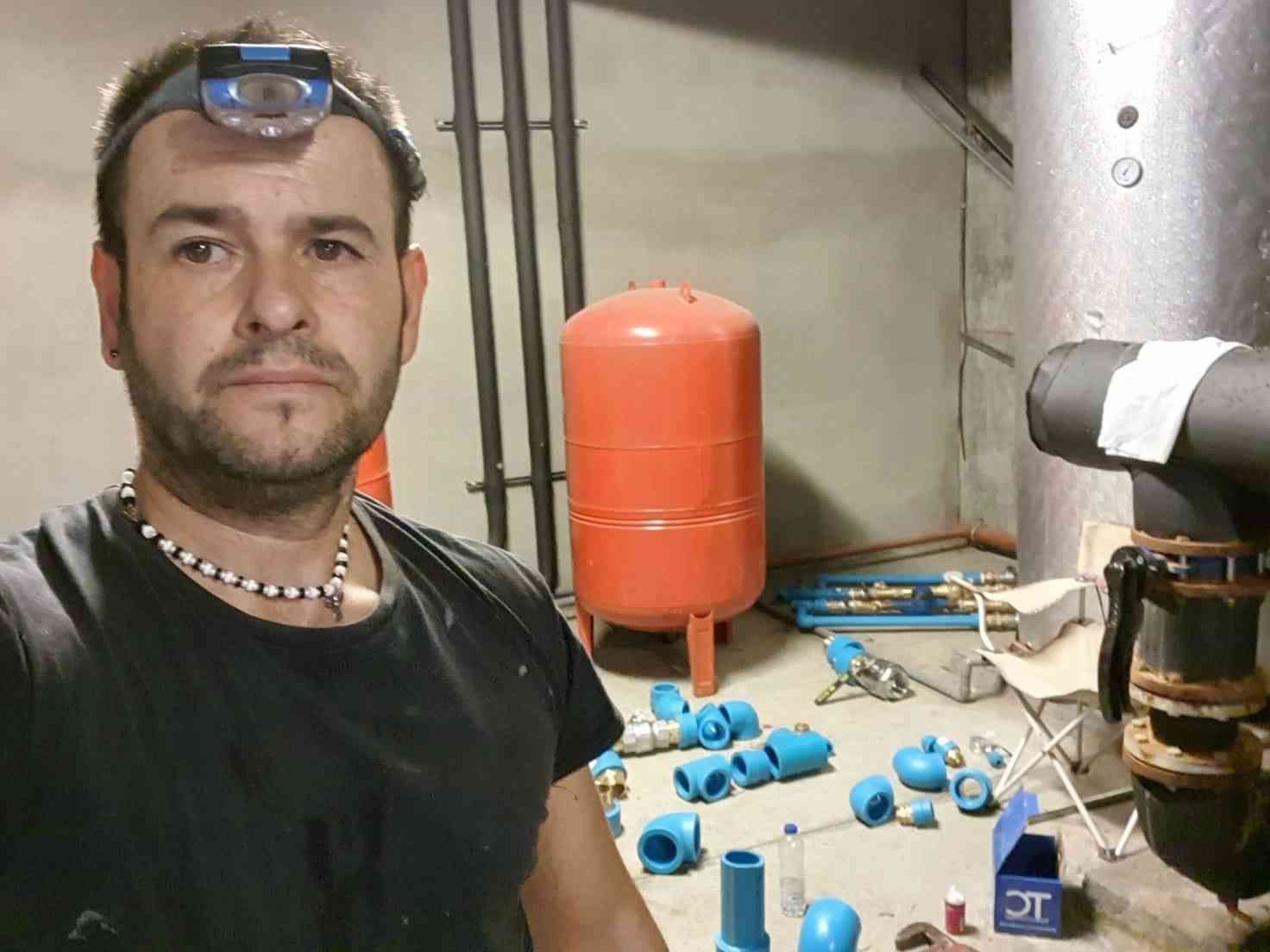 Hombre con linterna frontal en el lavadero, con herramientas de fontanería desperdigadas. Tanque naranja, tuberías y accesorios.